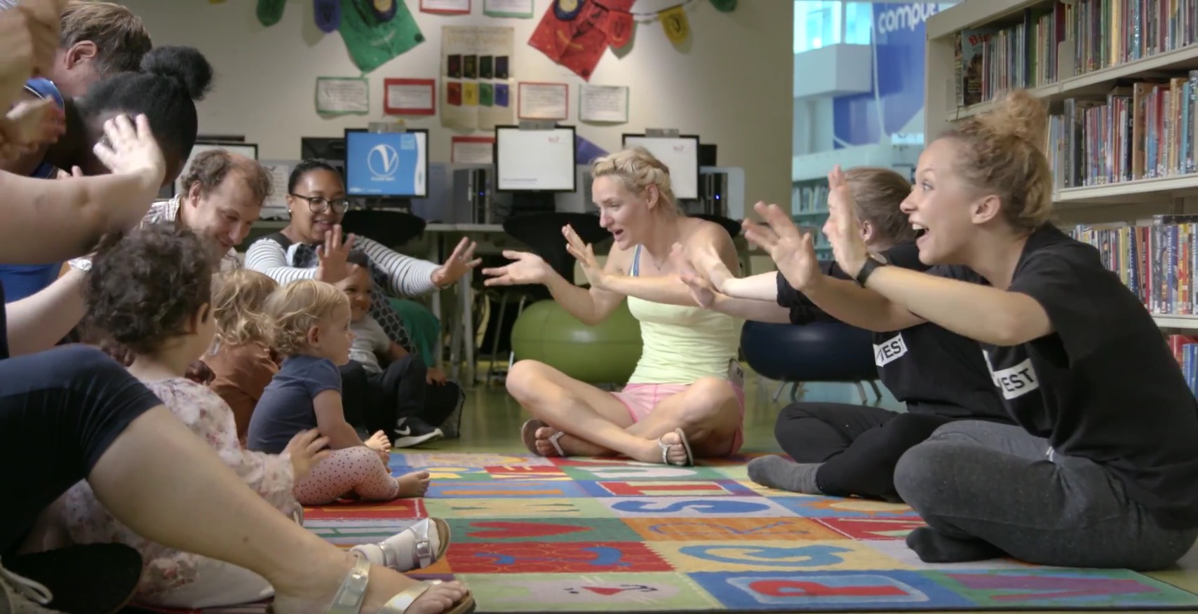 A photograph of a Dancing Books session - two leaders in black t-shirts face a group of toddlers and carers. All are looking excited and waving their hands in front of them.